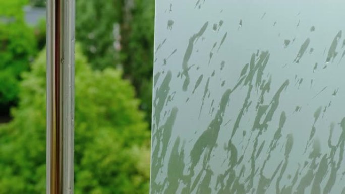 雨水落在绿色的哑光窗玻璃上，旁边是一根闪亮的金属杆，上面有水滴，背景是绿树。从公寓阳台上看，城市里有