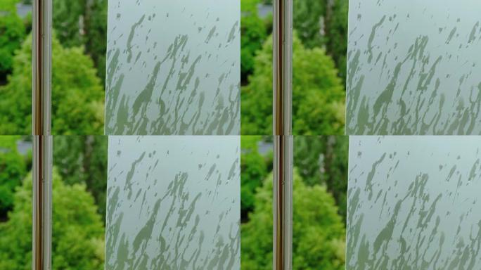 雨水落在绿色的哑光窗玻璃上，旁边是一根闪亮的金属杆，上面有水滴，背景是绿树。从公寓阳台上看，城市里有