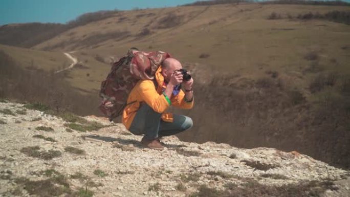 一个穿着黄色夹克，戴着眼镜的男人站在山上，欣赏着风景，用单反相机拍着照片。