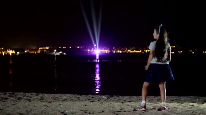 晚上，一个穿着闪亮裙子的少女在跳舞，从后面看。面对水面上发光城市的灯光