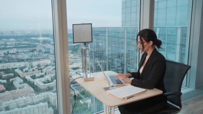 女商人在摩天大楼高楼层的现代办公室工作。迷人的女孩坐在大窗户旁的桌子旁，在笔记本电脑上工作。