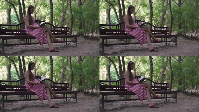 一个怀孕的女孩坐在公园的长椅上。一个穿着条纹白红色连衣裙的女孩热情地看书。