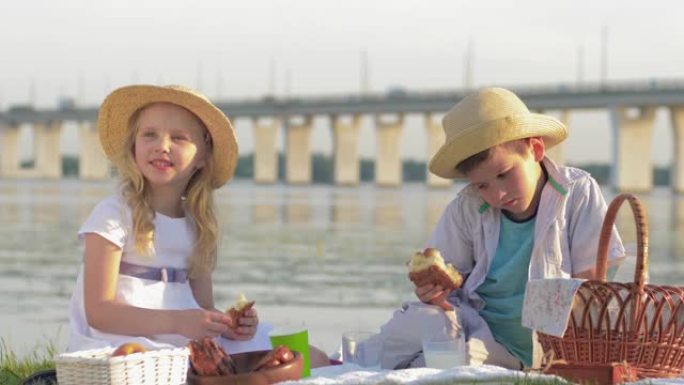 野餐的小朋友，饥饿的孩子男孩和女孩在桥背景下的河边自然野餐时吃甜面包和喝牛奶