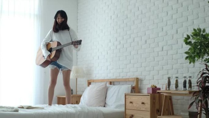 一名年轻女子在自然光的白色卧室的床上弹吉他，穿着白色和短牛仔裤的休闲装
