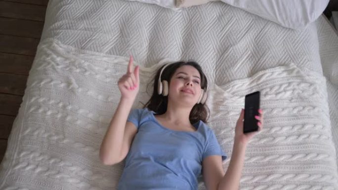 开朗的女性戴着耳机摔倒在床上，一边在家放松，一边听手机歌曲，一边听音乐