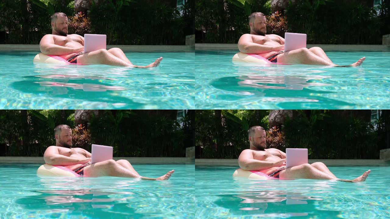 在游泳池的水中有一个充气环的男性在电脑上工作。粉色笔记本电脑游泳圈里有趣的胖子。自由职业概念