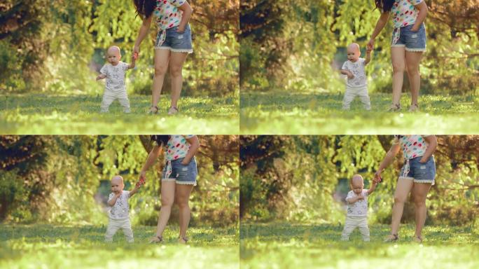 漂亮的宝贝站在绿色的草坪上，夏天在公园散步。小儿子牵着母亲的手。快乐的母亲。