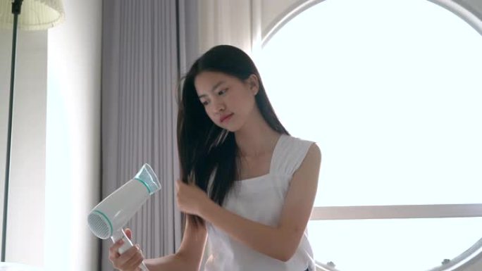 年轻漂亮的亚洲女人早上在酒店房间洗完澡后在床上用吹风机。