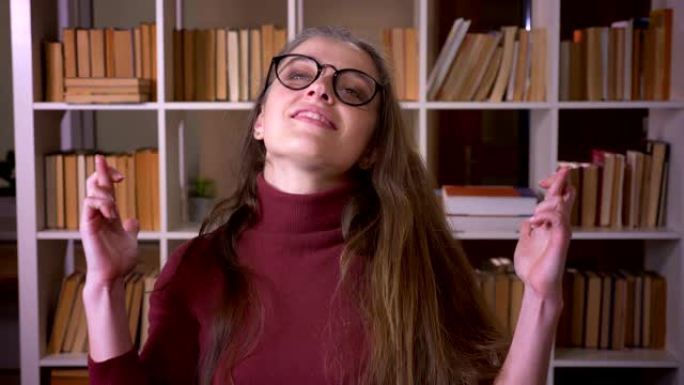 戴着眼镜的年轻高加索女学生的特写肖像，她的手指与希望交叉，看着室内大学图书馆的相机