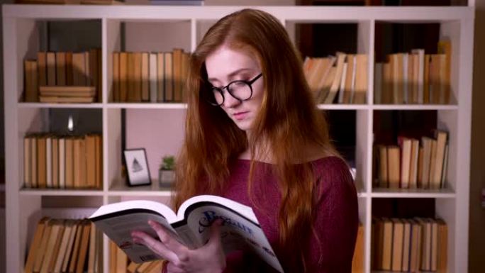戴着眼镜的年轻红发迷人白人女学生的特写肖像看着相机，在大学图书馆微笑着看书