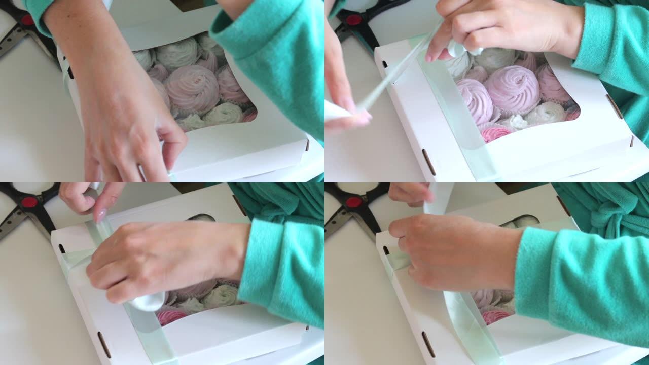 一个女人正在用绿松石辫子扎一盒棉花糖。不同颜色的棉花糖，通过盒子中的透明窗口可见。