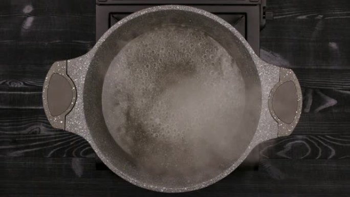 水在带有石材或大理石不粘涂层的金属锅中沸腾。平底锅放在煤气炉上，背景为黑色木桌。顶视图