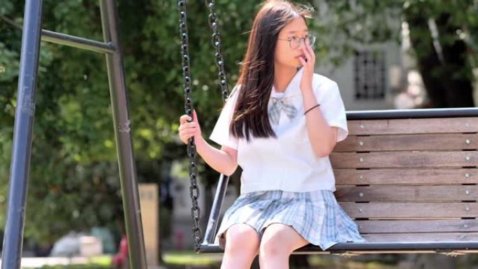 可爱的中国年轻女子，穿着日本学生风格的连衣裙，在阳光明媚的日子里，在公园的绳凳秋千上摇曳，戴着眼镜的