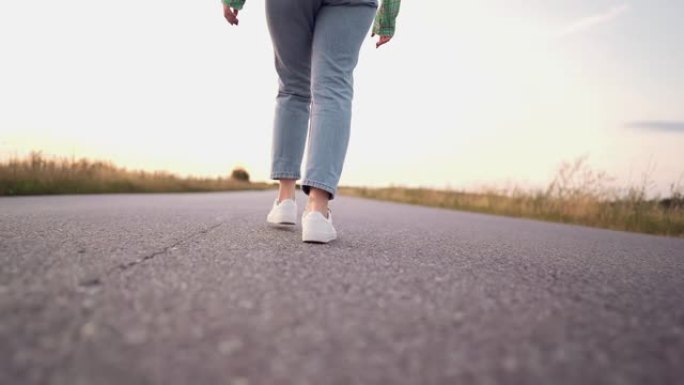 穿着牛仔布和新潮鞋的年轻女性在沥青光滑的道路上行走。现代运动鞋的概念，旅行，徒步旅行。后镜头。
