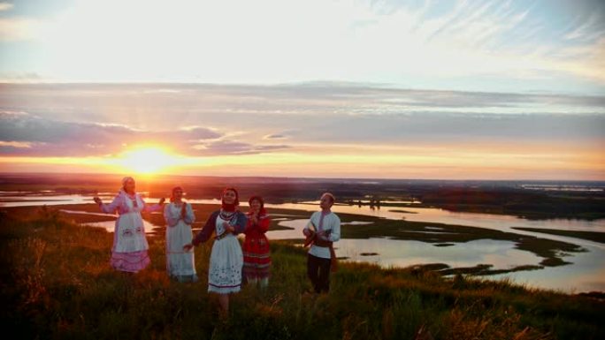 穿着俄罗斯传统服装的年轻人站在美丽的日落和唱歌的背景下。