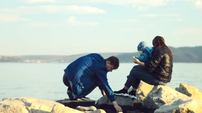 男人和家人坐在沙滩上的大石头上，向水中扔石头。女人怀里抱着一个小儿子。后视摄像头