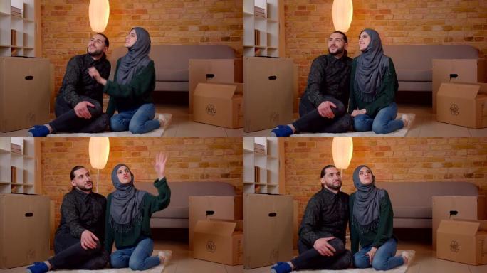 年轻开朗的穆斯林夫妇的特写镜头坐在新买的公寓里盒子旁边的地板上，微笑着幸福地梦想着如何为房间布置家具