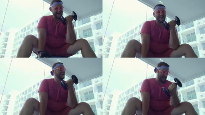 穿着粉红色眼镜和粉红色t恤的有趣胖胖的男性在健身房的一个健康球上与哑铃订婚，描绘了一个女孩