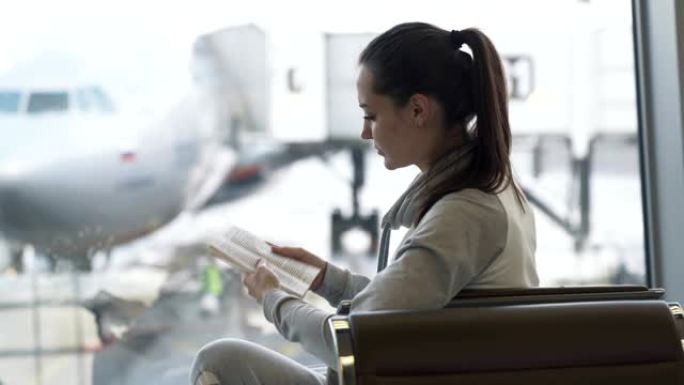 迷人女孩的侧视图坐在机场休息室，在窗户上阅读飞机背景的书
