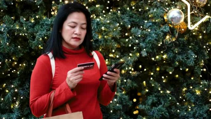 亚洲妇女在新年街市场交易会上使用信用卡在线购物，在圣诞树装饰上使用灯串。
