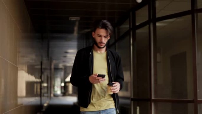 大胡子的年轻人手里拿着外卖咖啡杯走过隧道，发短信或用手机浏览互联网。男人在白天抬头沉思