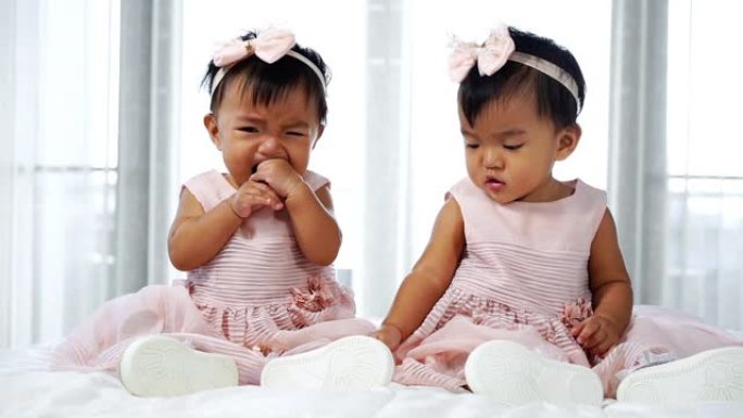 两个穿着粉红色连衣裙的双胞胎婴儿在床上，一个看，一个哭