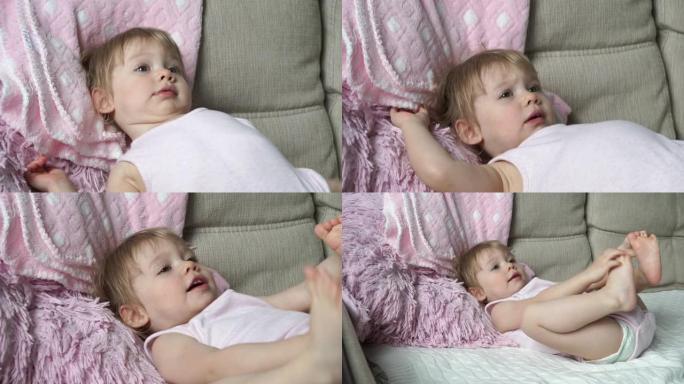 穿着粉红色衣服的2岁小女孩的肖像躺在床上，用脚玩耍，微笑着