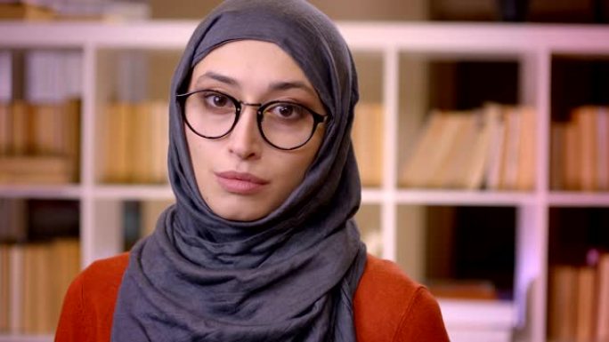 盖头的年轻有吸引力的穆斯林成功女学生的特写镜头固定眼镜看着站在图书馆室内的相机