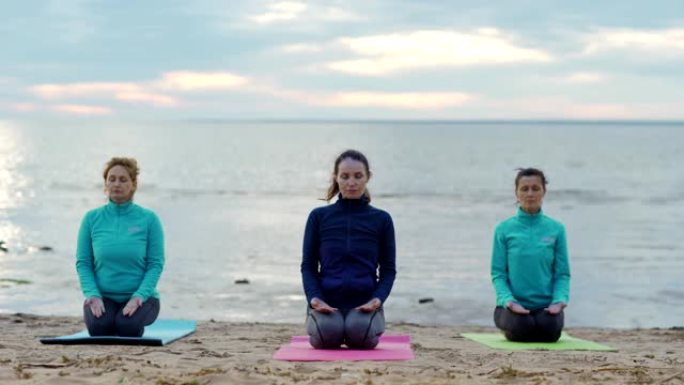 三个成熟女性在海滩上练习瑜伽。妇女跪在海边的垫子上，闭着眼睛冥想