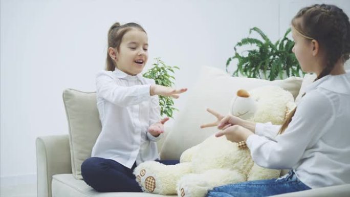 两个漂亮的女孩坐在沙发上，玩石头剪刀布，决定谁将与白色柔软的熊一起玩。