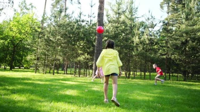 在夏季公园的绿色草坪上，三个孩子用红球玩轰击游戏。女孩用慢动作投球试图击中对手