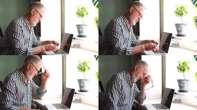成熟的大胡子男人在家用笔记本电脑工作。坐在窗户附近的桌子上