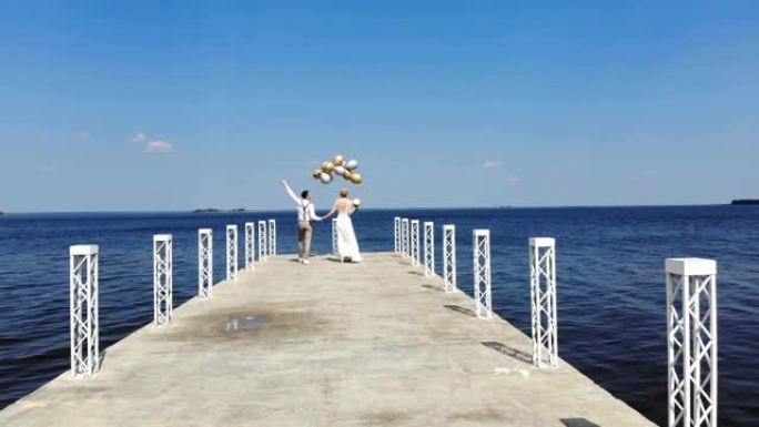 年轻漂亮的一对新婚夫妇走在美丽的码头上，新郎手里拿着气球，面面相视，面带微笑，靠着蔚蓝的大海和天空。