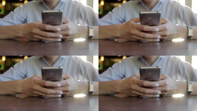 年轻的女商人穿着蓝色衬衫和金色手表，坐在餐厅里用智能手机打字，双手放在桌子上。