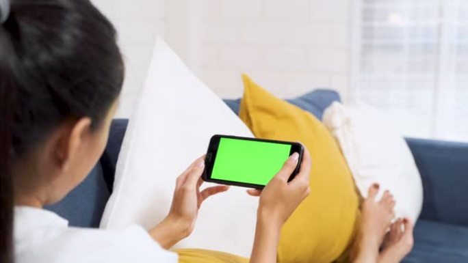 亚洲妇女躺在沙发上，在家中观看手机上的视频绿屏，令人不寒而栗。数字时代的生活方式。