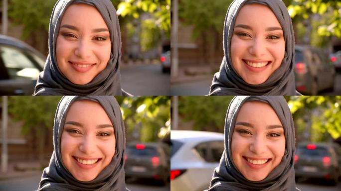 戴着头巾的穆斯林学生的肖像站在绿色城市街道上欢快地对着镜头微笑。
