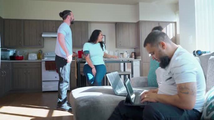 这位年轻的大胡子男子在共用出租公寓的客厅的沙发上使用笔记本电脑和平板电脑，当时他的室友在厨房的背景下