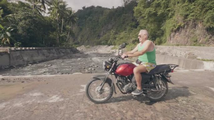 暑假期间，老人在乡村路上骑摩托车。旅行者男子在亚洲村的河桥上骑摩托车。摩托旅行。