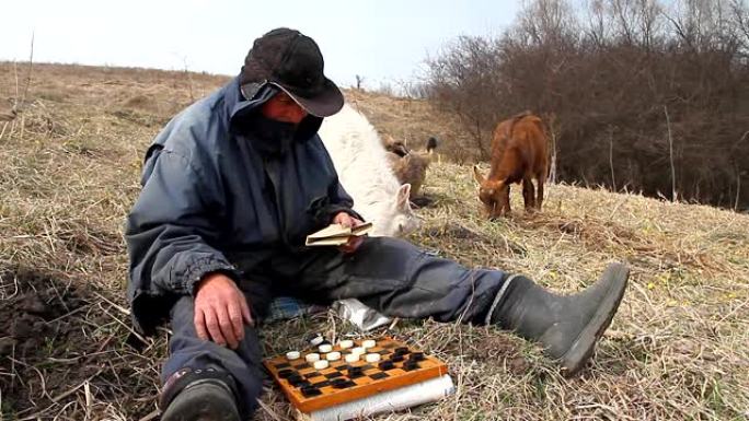 一位穿着凌乱衣服的老人坐在山上玩跳棋，在大自然枯萎的背景下放牧一群自己的山羊