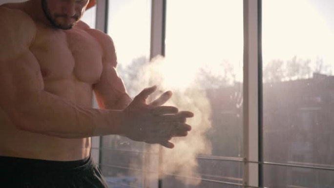 肌肉发达的男子用白色粉笔粉拍打着双手，准备在体育中心进行塑身力量训练的慢动作