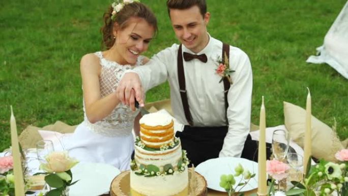 婚礼当天，新郎和新娘坐在草地上切蛋糕，在梳妆树和白色装饰有桌子和蛋糕的帐篷之间。