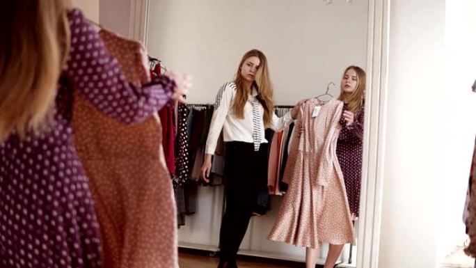 两个金发双胞胎姐妹正在一起购物。姐姐尝试在镜子前穿时髦的衣服，姐姐愿意尝试另一件衣服。站在衣架附近的