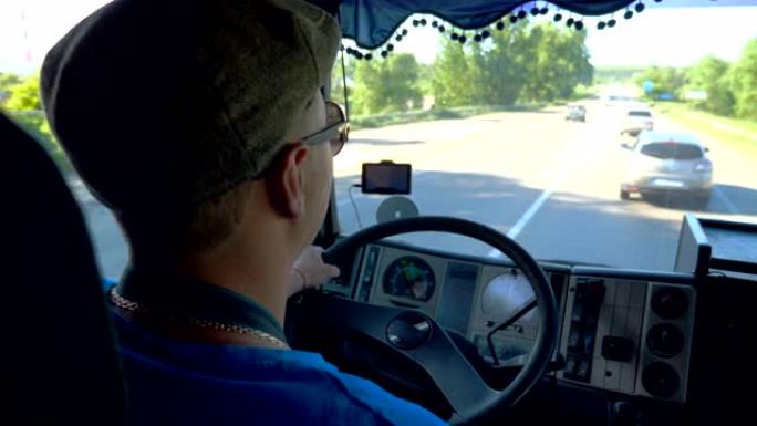 晴天，无法识别的卡车司机在县道上行驶。戴着帽子和墨镜的男子控制着他的卡车专心观看道路。从机舱内查看交