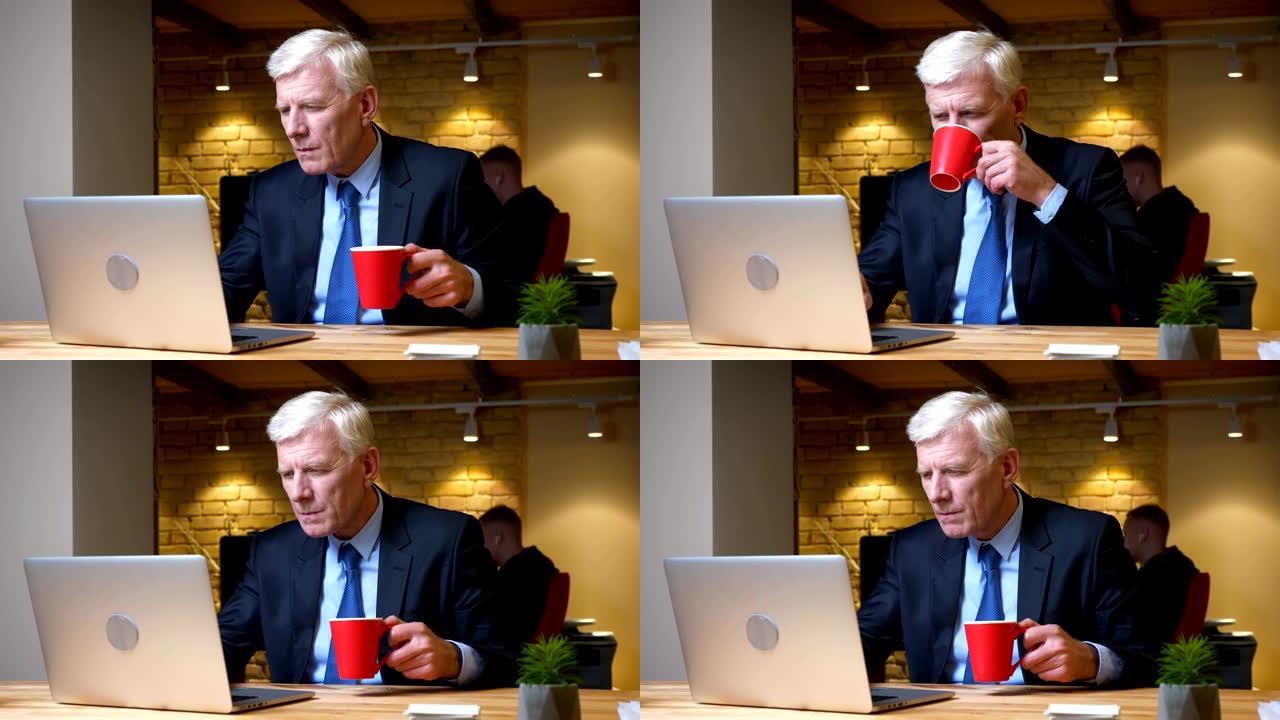 老高加索商人使用笔记本电脑和喝咖啡的特写镜头在工作场所的办公室里忙碌而专注于室内