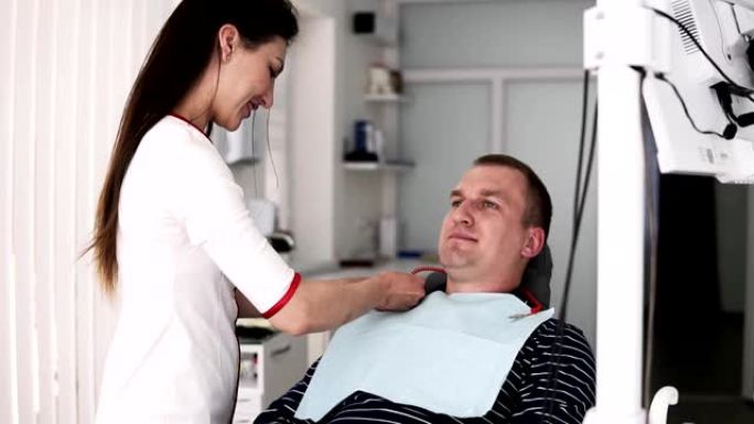长毛女牙医在口腔诊疗室的背景下为男性病人准备牙齿治疗。医生在一个坐在牙医椅上的年轻人的胸膛上盖了一张