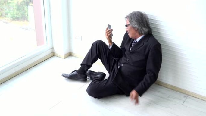 端庄的老商人坐在地板上，在窗户旁边失望地看着智能手机