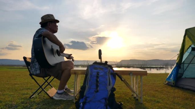 亚洲老人的场景慢动作正在弹吉他，在暑假期间欣赏湖边的景色。他很开心，在假期放松的时候也很开心。户外自