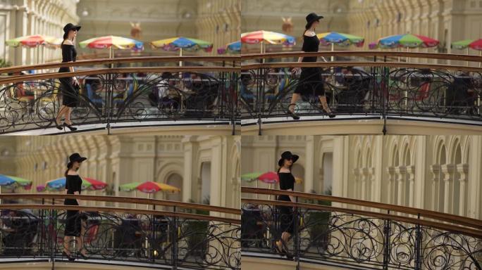 穿着黑色连衣裙，太阳镜和帽子的可爱时尚女孩在商场的桥上。经典优雅的时尚女性。慢动作