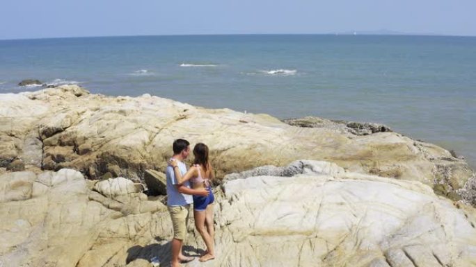 一对恩爱的夫妇沿着沙滩的金色沙滩散步，在石头上行走，互相帮助并自拍。从上方观看