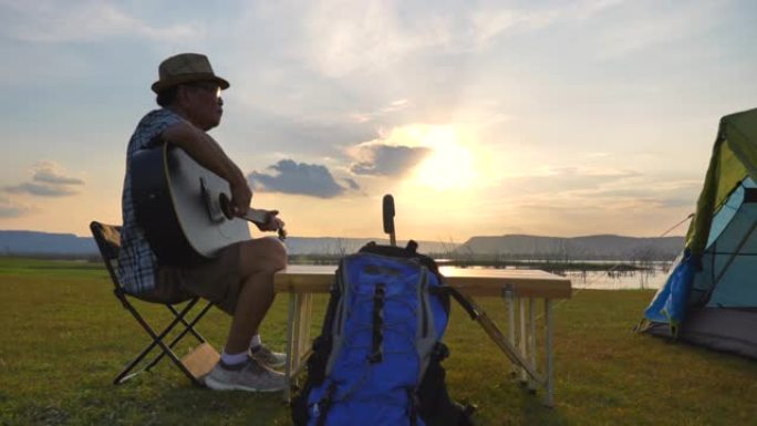 亚洲高级男性在唱歌时弹吉他的慢动作场景令人放松，并在暑假期间欣赏湖边的景色。他很开心，在假期、放松的